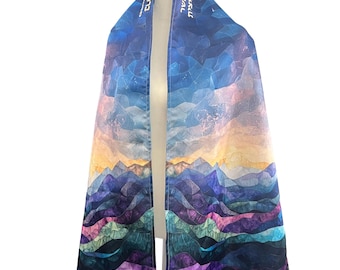 May include: A tallit prayer shawl featuring a vibrant, multi-colored landscape print of mountains and sky in shades of deep blue, purple, orange, and teal. The atarah, or neckband, displays white Hebrew text against a dark blue background.