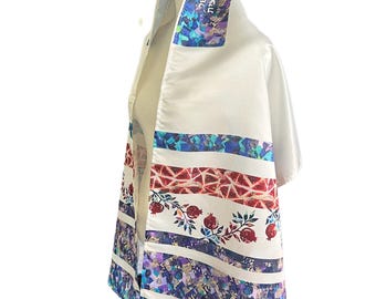 Può includere: Uno scialle da preghiera tallit bianco con un'atarah decorativa con testo in ebraico. Il tessuto presenta fasce orizzontali con motivi a mosaico colorati nelle tonalità del blu, viola e rosso, intervallate da illustrazioni di melograni.