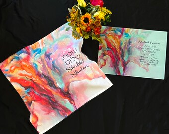 Può includere: Un design astratto colorato stampato su una tovaglia da Shabbat in tessuto bianco con le parole ebraiche "Shabbat Shalom" in lettere nere. Un tagliere di vetro abbinato con lo stesso design e le parole ebraiche "Shabbat Shalom" e la traduzione inglese "Blessed are You, Lord our God who brings forth bread from the earth" in lettere nere.