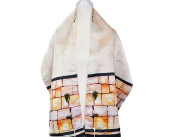 May include: A cream-colored tallit, a Jewish prayer shawl, featuring a watercolor design of the Western Wall. The design includes tan and brown bricks, green plants, and black stripes. The shawl has white tassels and is draped over a white stand.