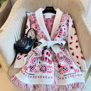 May include: A white cardigan with a red and black paisley pattern and fringe trim. The cardigan is tied with a white bow and is displayed on a beige armchair. A black handbag sits to the left.