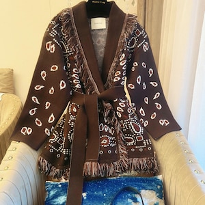 May include: A brown patterned cardigan with a matching belt and fringe detailing. The open-front sweater features a geometric design in white and red. The cardigan is displayed on a chair with a blue sequined pillow.