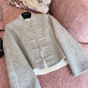 May include: A light gray jacket with a mandarin collar and flared sleeves. The jacket features four decorative frog closures with small purple buttons. The jacket is displayed on a hanger against a pink background.