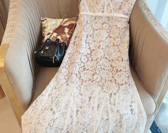 May include: A white lace dress with short sleeves and a square neckline. The dress features a fitted bodice, a defined waist, and a flowing skirt. A black purse with a chain strap rests on the chair.