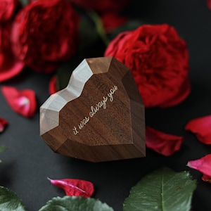 Op de afbeelding: Een houten hartvormige doos met een geometrisch ontwerp. De doos is gegraveerd met de woorden "it was always you". De doos is omringd door rode rozenblaadjes en groene bladeren.