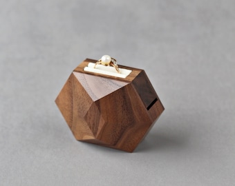 May include: A hexagonal wooden ring box with a geometric design. The box is made of walnut wood and has a white velvet interior. A gold ring with a pearl is displayed in the box.