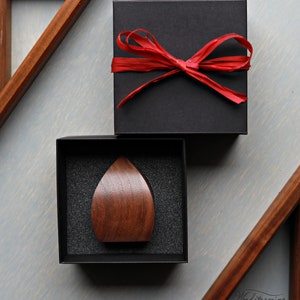 May include: A dark brown wooden teardrop-shaped object is displayed in a black box with a black foam interior. The box is sitting on a light gray surface. A black box with a red ribbon is sitting on top of the box.