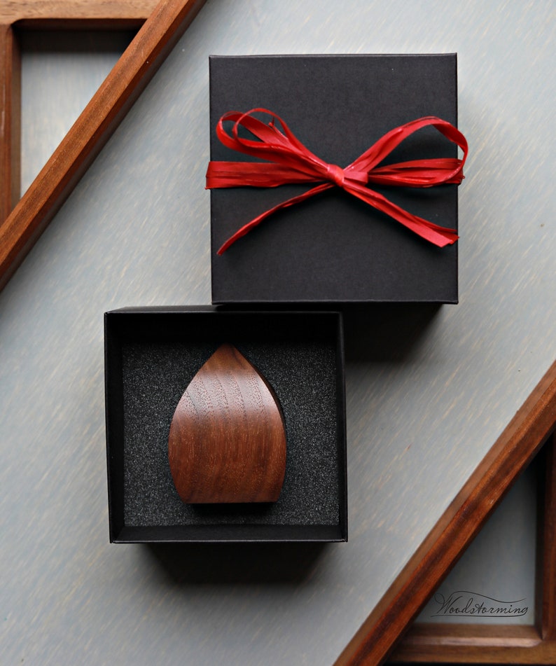 May include: A dark brown wooden teardrop-shaped object is displayed in a black box with a black foam interior. The box is sitting on a light gray surface. A black box with a red ribbon is sitting on top of the box.