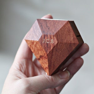 Puede incluir: Una caja de anillo de madera con forma de diamante con las iniciales "J.C.B." grabadas en la parte delantera.