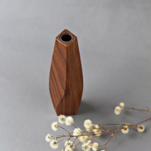 May include: A wooden vase with a geometric design. The vase is made of dark brown wood and has a round opening at the top. The vase is sitting on a gray surface.