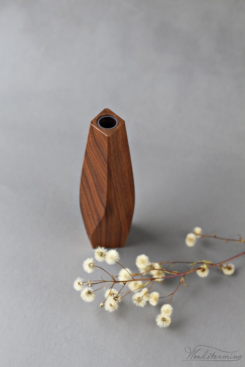 May include: A wooden vase with a geometric design. The vase is made of dark brown wood and has a round opening at the top. The vase is sitting on a gray surface.