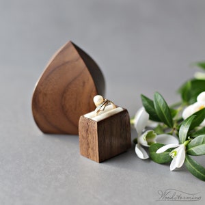 May include: A wooden ring box with a heart-shaped lid and a smaller square box inside. The ring box is made of walnut wood and has a natural finish. The smaller box is also made of walnut wood and has a white velvet lining. A ring with a pearl and diamonds is sitting in the smaller box.