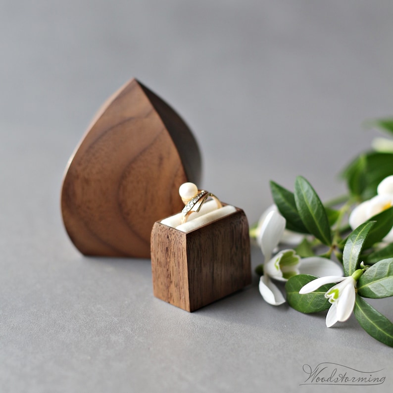May include: A wooden ring box with a heart-shaped lid and a smaller square box inside. The ring box is made of walnut wood and has a natural finish. The smaller box is also made of walnut wood and has a white velvet lining. A ring with a pearl and diamonds is sitting in the smaller box.