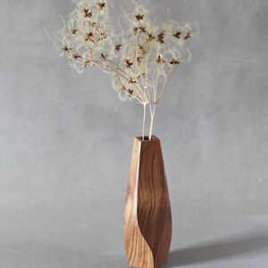 May include: A wooden vase with a unique, curved design, holding dried flowers with fluffy seed heads. The vase is made of dark wood with a natural grain pattern.
