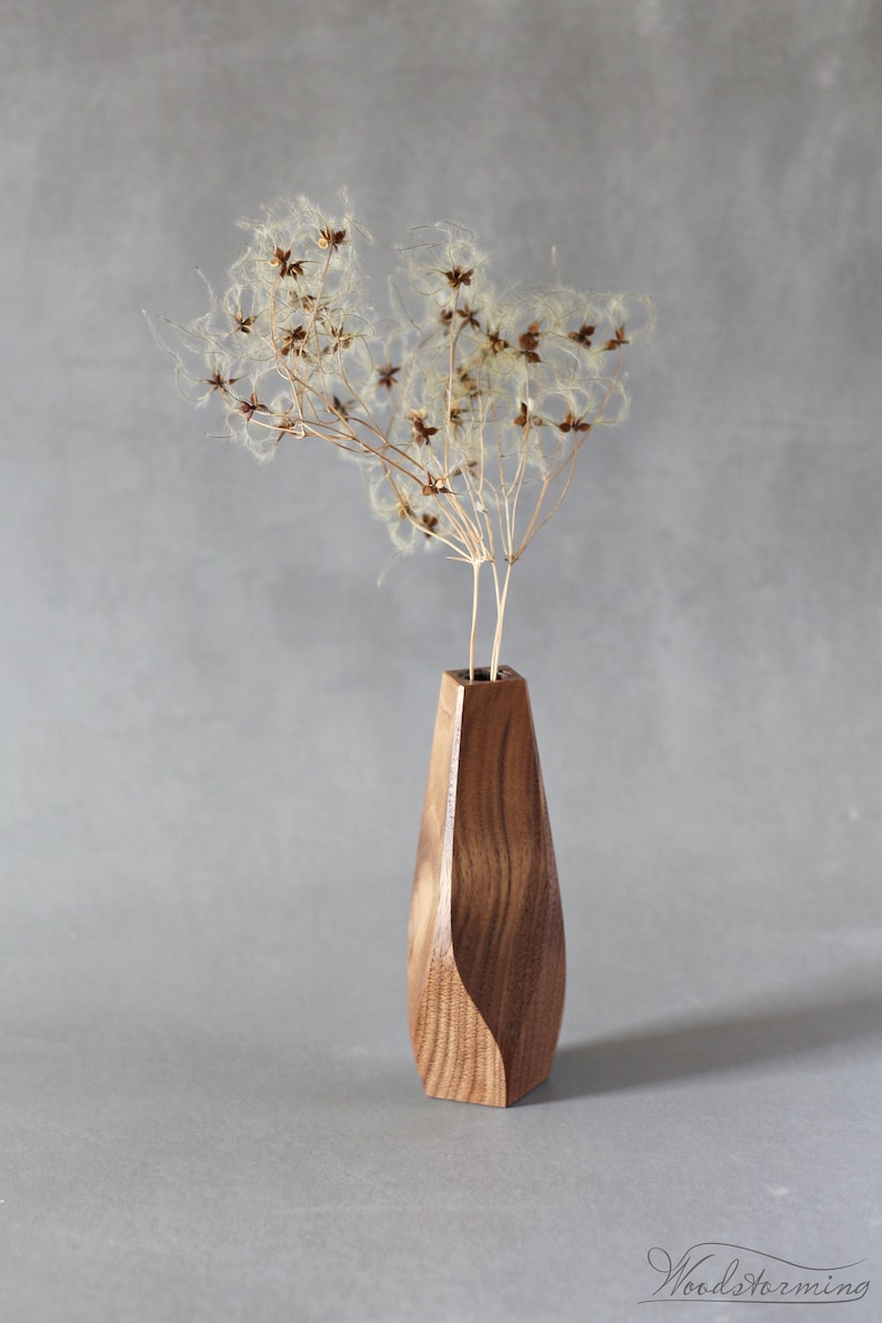 May include: A wooden vase with a unique, curved design, holding dried flowers with fluffy seed heads. The vase is made of dark wood with a natural grain pattern.
