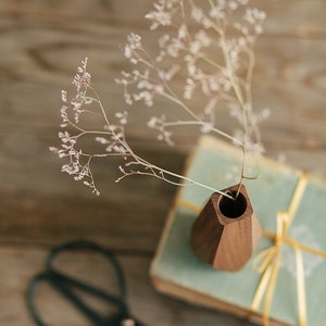 May include: A wooden vase with a geometric design holds a bouquet of dried white flowers. The vase is sitting on top of a stack of books with a gold ribbon.