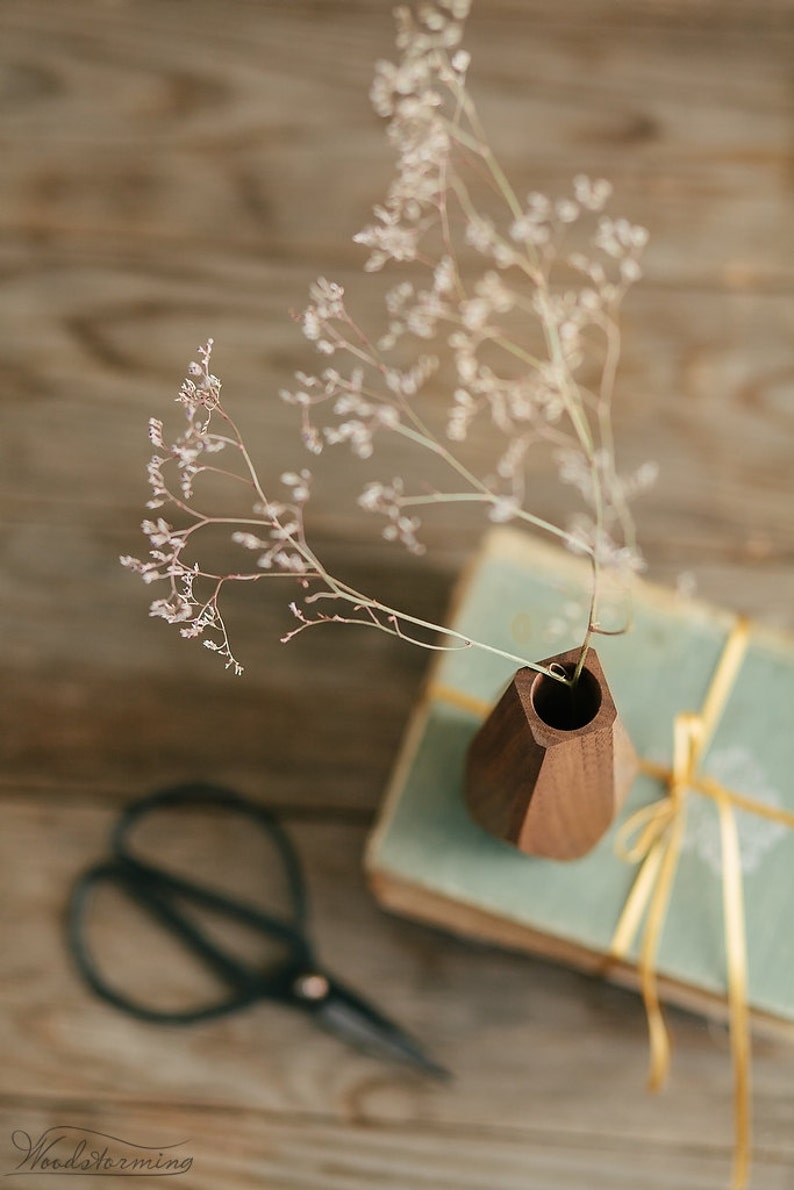 May include: A wooden vase with a geometric design holds a bouquet of dried white flowers. The vase is sitting on top of a stack of books with a gold ribbon.