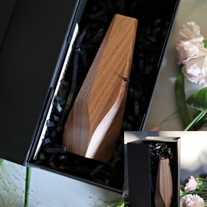 May include: A wooden vase with a geometric design, in a black box with black shredded paper. The vase is made of walnut wood and has a natural finish.