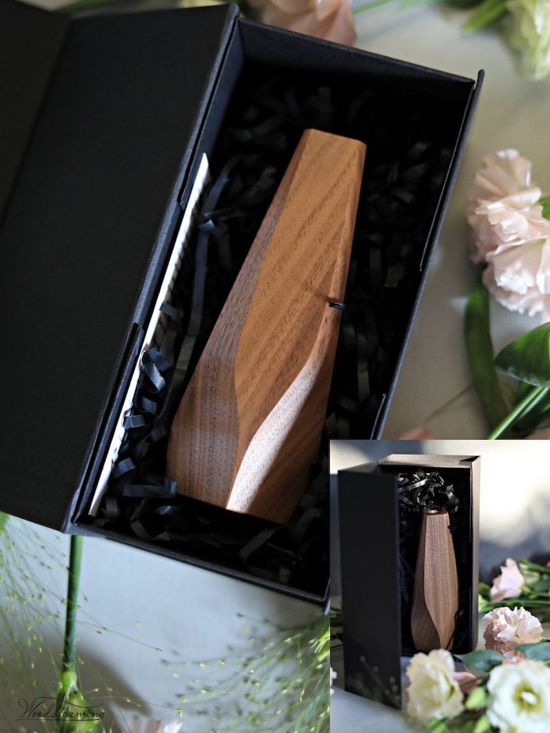 May include: A wooden vase with a geometric design, in a black box with black shredded paper. The vase is made of walnut wood and has a natural finish.