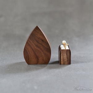 May include: Two wooden ring boxes, one shaped like a teardrop and the other a rectangle, both made of dark walnut wood. A gold ring with a white stone is displayed in the rectangular box.