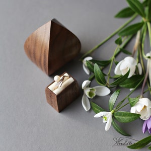 May include: A wooden ring box with a ring inside. The box is made of walnut wood and has a simple, modern design. The ring is a gold band with a pearl and small diamonds. The box is sitting on a gray surface with white flowers.