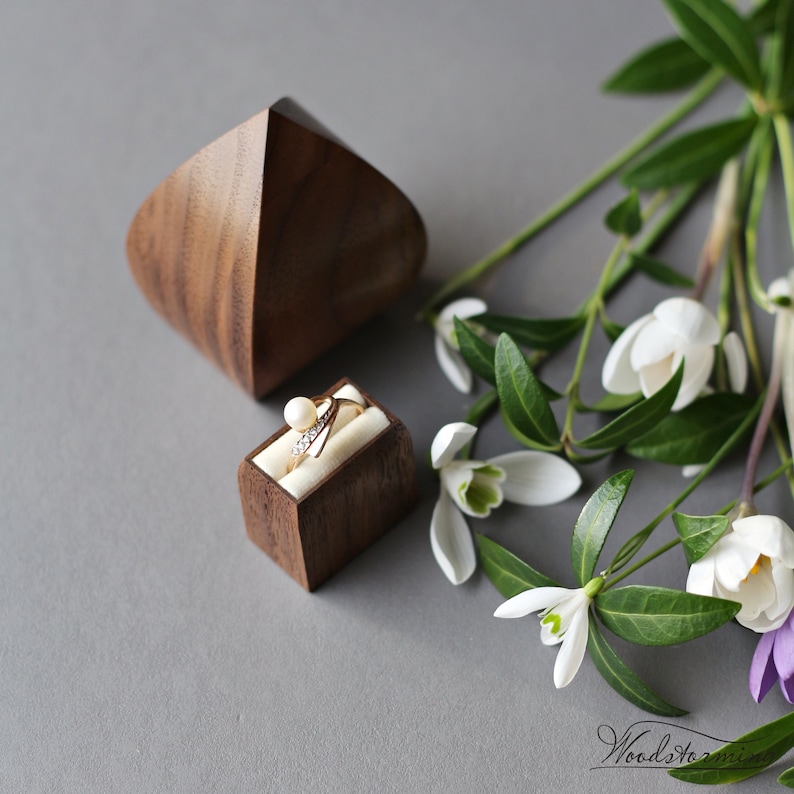 May include: A wooden ring box with a ring inside. The box is made of walnut wood and has a simple, modern design. The ring is a gold band with a pearl and small diamonds. The box is sitting on a gray surface with white flowers.