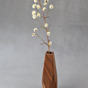May include: A wooden vase with a geometric shape and a dark brown wood grain. The vase is filled with a single stem of dried white flowers.