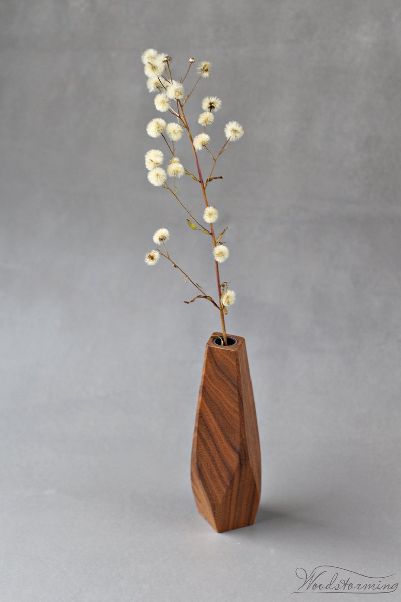 May include: A wooden vase with a geometric shape and a dark brown wood grain. The vase is filled with a single stem of dried white flowers.