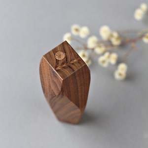 May include: A small, geometric wooden vase made of walnut wood. The vase has a unique, faceted design and a circular, wooden top with a carved design.