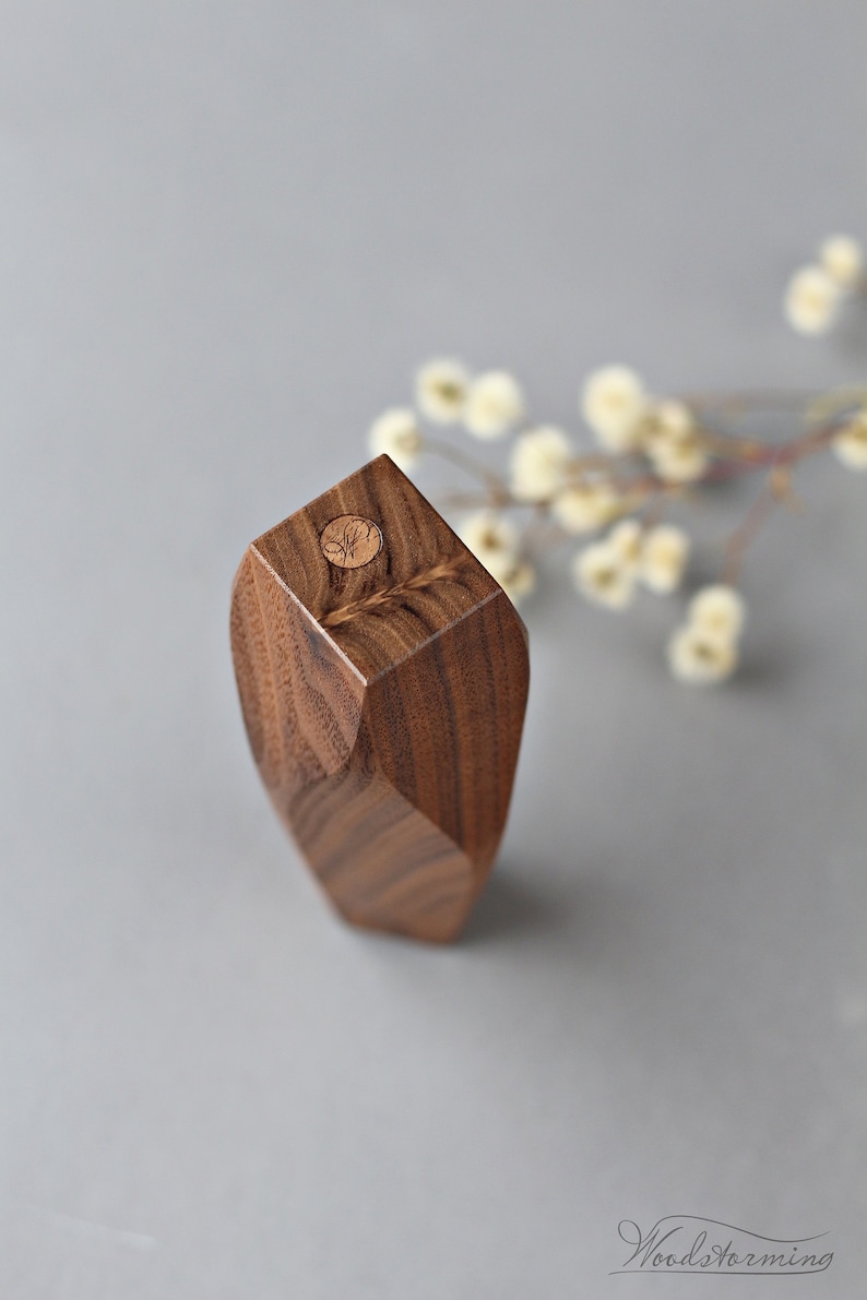 May include: A small, geometric wooden vase made of walnut wood. The vase has a unique, faceted design and a circular, wooden top with a carved design.