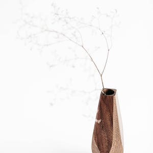 May include: A dark brown wooden vase with a geometric design. The vase is empty and has a small opening at the top. A dried flower stem is in the vase.