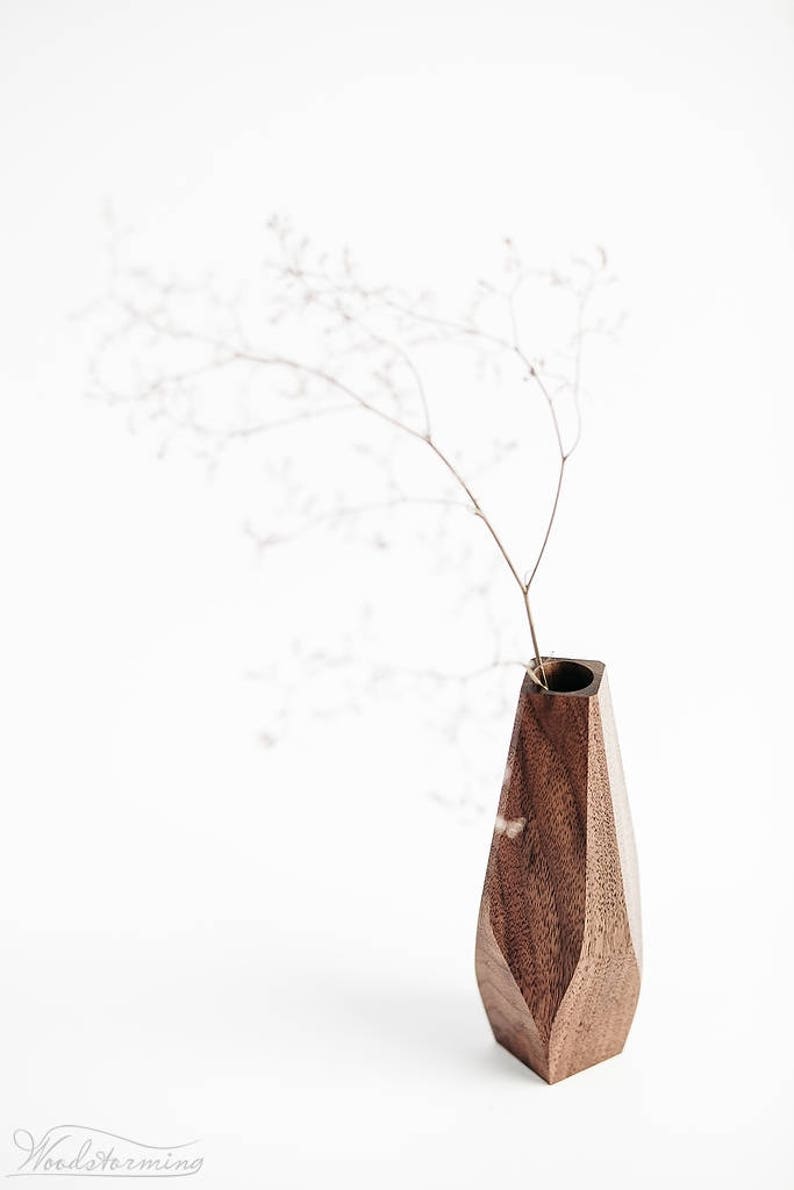 May include: A dark brown wooden vase with a geometric design. The vase is empty and has a small opening at the top. A dried flower stem is in the vase.