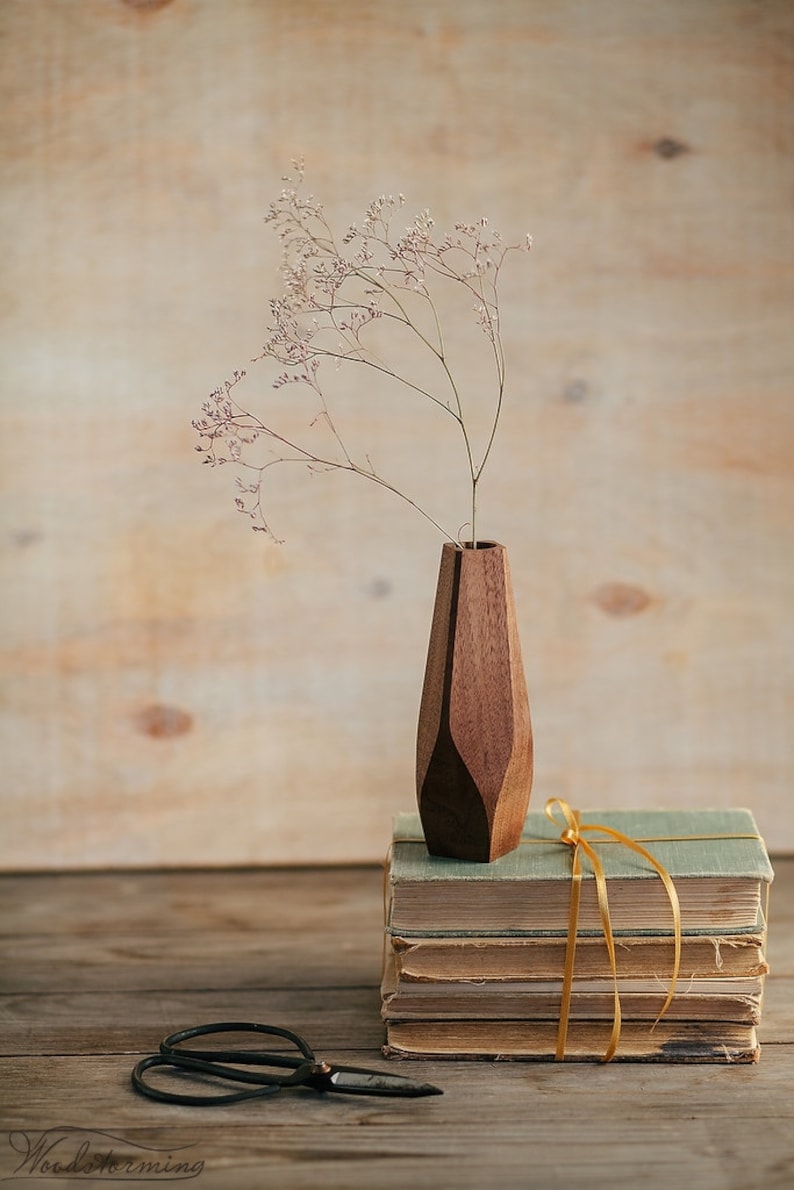 May include: A wooden vase with a geometric design holds a bouquet of dried flowers. The vase is sitting on top of a stack of old books tied together with a yellow ribbon.