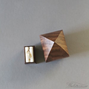 May include: A wooden ring box with a pyramid-shaped lid. The box is made of dark wood and has a natural wood grain. The lid is open and a gold ring with a diamond is inside.