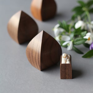 May include: Three wooden ring boxes in the shape of a teardrop, with one box containing a gold ring with a pearl and diamonds. The boxes are on a gray surface with a sprig of white flowers.