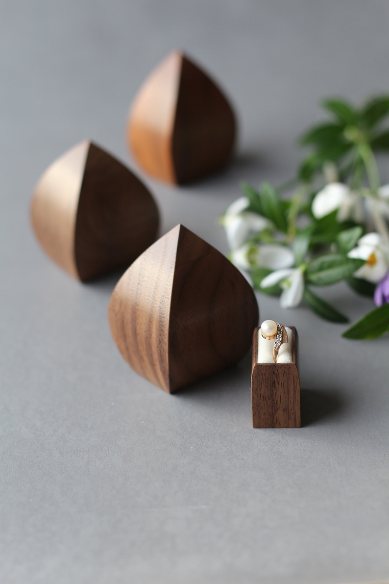 May include: Three wooden ring boxes in the shape of a teardrop, with one box containing a gold ring with a pearl and diamonds. The boxes are on a gray surface with a sprig of white flowers.