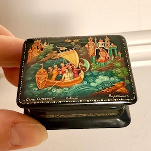 Puede incluir: Una pequeña caja rectangular de laca negra con una escena pintada a mano de un barco con varias figuras en el mar y edificios ornamentados al fondo. La tapa incluye texto en cirílico y un borde decorativo de color dorado en los lados.