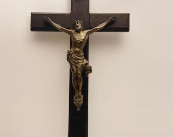 May include: A dark brown wooden crucifix featuring a metallic bronze-toned figure of Jesus Christ attached to the center. Above the figure, a small silver-colored plaque is mounted to the wood, displaying the engraved letters INRI.