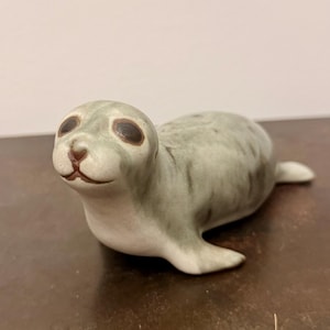 Puede incluir: Una figura de cerámica de una cría de foca con un cuerpo liso de color gris claro y manchas moteadas de color gris oscuro. La foca tiene grandes ojos de color marrón oscuro, una nariz pequeña y una línea de boca sutil en reposo.
