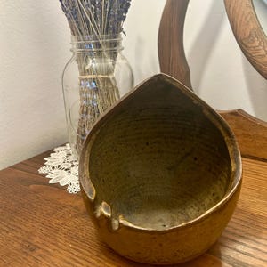 May include: A handmade, ceramic bowl with a unique, organic shape and a textured surface. The bowl has a warm, earthy color palette, with shades of brown and gold. A glass jar with dried lavender is in the background.