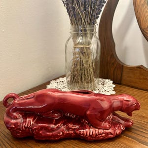 Peut inclure: Une figurine de panthère en céramique rouge brillant, couchée, la tête levée. La sculpture repose sur une base texturée. Un bocal en verre avec de la lavande séchée est en arrière-plan, ajoutant une touche de décoration naturelle.
