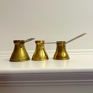 May include: Three brass coffee pots with a flared body and long handle. The pots are arranged in a row, displaying their shiny, golden colour. The background is a plain white surface.