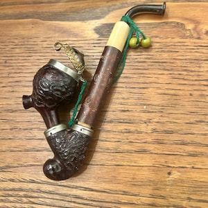 May include: A dark brown tobacco pipe with a curved stem and a decorative chain. The pipe has a textured bowl and a light-colored section near the mouthpiece. Green string and small yellow beads are attached.