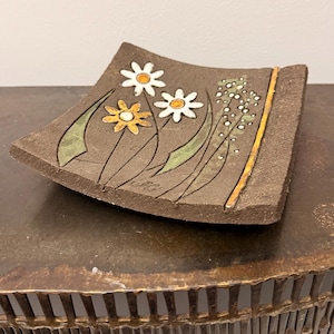 May include: A square, dark brown ceramic dish with a textured surface featuring a hand-painted design of two white daisies, one yellow flower, green leaves, and a cluster of small white dots, accented by a vertical orange line on the right side.