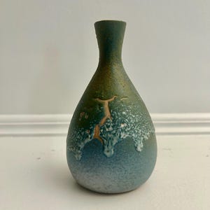 May include: A small, handcrafted ceramic vase with a unique, textured surface. The vase features a gradient of colours, transitioning from a deep green at the top to a blue hue at the base. A crack-like design adds a touch of rustic charm.