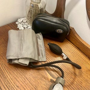 Puede incluir: Un tensiómetro vintage con un manguito gris, una pera negra y un medidor. Junto al tensiómetro hay un estuche de cuero negro con cremallera plateada y un pequeño emblema. Un tarro de cristal con lavanda seca y un tapete de encaje blanco están en el fondo.