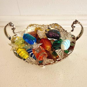 May include: A silver-toned metal bowl with ornate handles and a scalloped rim, filled with various glass candy-shaped ornaments. The glass pieces feature diverse colours including blue, orange, yellow, green, and black with glittery accents.