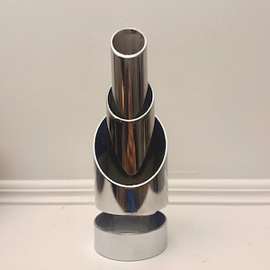 May include: A polished chrome metal sculpture featuring three cylindrical tubes of varying heights arranged vertically. The tubes are cut at an angle at the top and are supported by a circular base, creating a tiered, reflective, and metallic structure.
