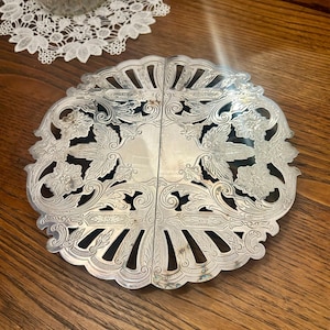 Op de afbeelding: Een sierlijke zilverkleurige metalen onderzetter met een bloemen- en krulontwerp. De onderzetter is opengewerkt, met een centraal rechthoekig gedeelte en scharnierende delen. Het ontwerp omvat bloemen, bladeren en gebogen lijnen.