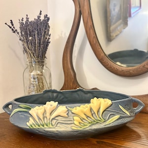 Peut inclure: Un bol en céramique bleu avec des motifs floraux jaunes en relief. Le bol a deux poignées et est posé sur une surface en bois. Un bocal en verre avec de la lavande séchée est en arrière-plan.